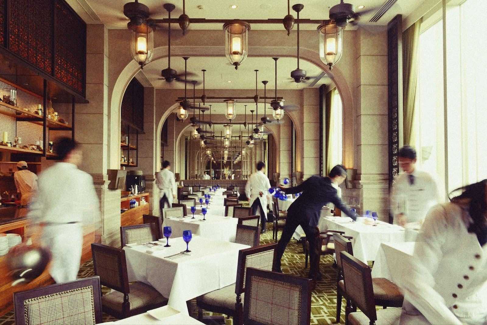 白いクロスがかかったテーブルが並ぶレストランでユニフォームを着用したスタッフが働いている様子