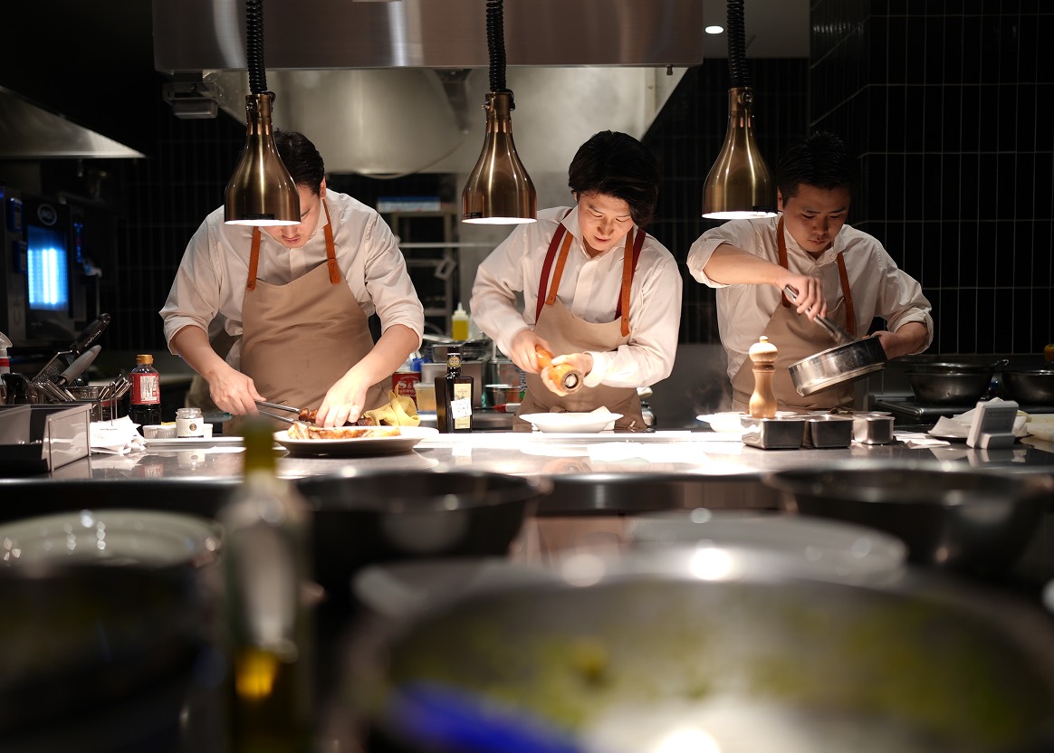 ベージュに赤の紐がついたエプロンを着用したレストランのキッチンスタッフ3名が働いている様子