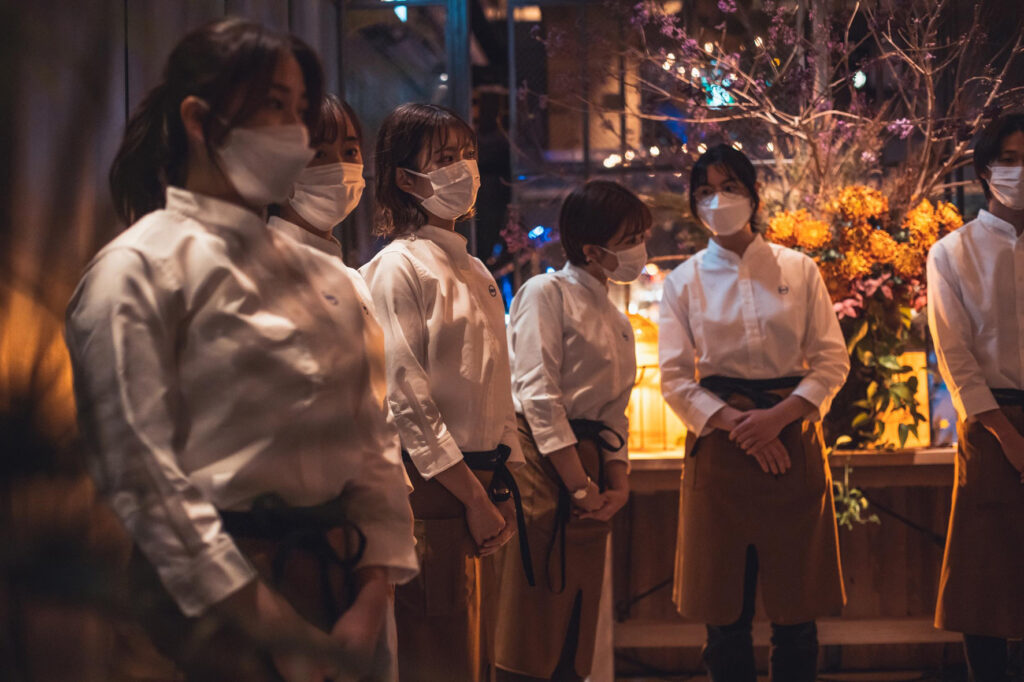 白いシャツにベージュのエプロンを着用したレストランスタッフが集合している様子